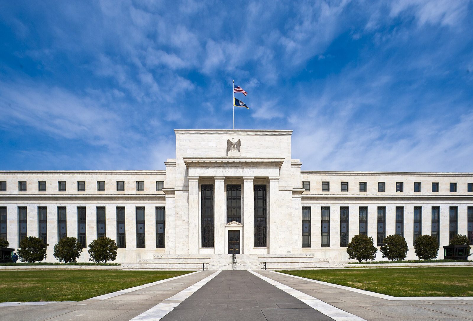 Front facade of the Marriner S. Eccles Federal Reserve Board building in Washington, DC, with the US and Federal Reserve flags flying against a blue sky
